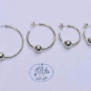 May include: Four silver hoop earrings arranged in descending order of size, each featuring a single polished silver bead that slides along the hoop. The earrings have standard post-back closures and are displayed with a small tree logo stamp.
