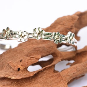 May include: A silver cuff bracelet featuring five evenly spaced flower motifs along the band. Each flower has six petals with a subtle green tint and a small, polished gold-tone bead at the center. The bracelet has a smooth, reflective finish.