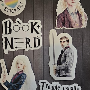 May include: A collection of stickers featuring characters and quotes from a fantasy book series. Includes a sticker with the text "Book Nerd" and another with the phrase "Trouble usually finds me". Other stickers depict character portraits and a heart-shaped sticker.