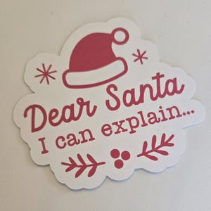 Può includere: Un adesivo bianco con bordo smerlato, con la scritta "Dear Santa I can explain..." in rosso. Un cappello di Babbo Natale rosso, fiocchi di neve e rametti di agrifoglio sono inclusi nel design.