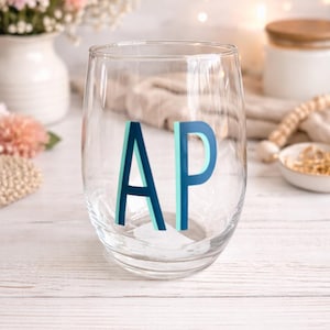 May include: A clear stemless wine glass with the letters "AP" in blue and teal. The glass sits on a white wooden surface, with flowers and a candle in the background. The glass is likely used for beverages.