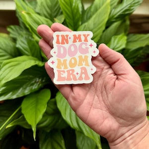 May include: A sticker with the text "IN MY DOG MOM ERA" in a retro font. The text is in shades of orange, pink, and peach, with small star accents. The sticker is held in a hand against a backdrop of green leaves.