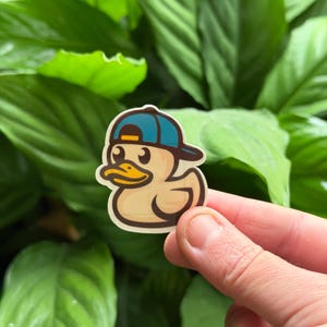 May include: A die-cut sticker featuring a cartoon yellow rubber duck wearing a blue baseball cap with a yellow brim. The duck has a dark brown outline, a bright yellow beak, and large black eyes, presented as a graphic illustration on a white background.