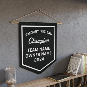 May include: Black pennant banner with white text that reads "FANTASY FOOTBALL Champion TEAM NAME OWNER NAME 2024". The banner is hanging from a wooden dowel with a string. The banner is hanging on a gray wall.
