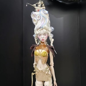 Puede incluir: Muñeca de arte detallada con tez pálida, elaborada peluca blanca y azul, y un barco en miniatura en la parte superior. La muñeca viste un corpiño dorado y marrón, con detalles a juego. La muñeca está de pie, con una expresión neutra.