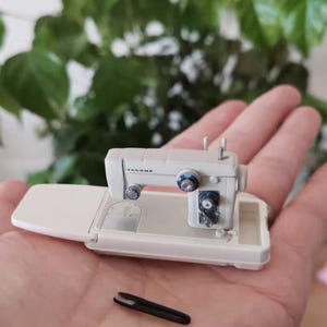 Może przedstawiać: Miniaturowa maszyna do szycia w kolorze złamanej bieli z niebieskimi i srebrnymi akcentami, spoczywająca na białej podstawie. Małe czarne nożyczki leżą poniżej. Maszyna trzymana jest w dłoni, z rozmytym zielonym tłem roślinnym.