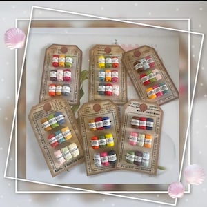 Puede incluir: Una colección de madejas de hilo en miniatura de varios colores, empaquetadas en etiquetas de estilo vintage. Cada etiqueta contiene varios pequeños paquetes de hilo, mostrando una gama de tonos, desde pasteles hasta colores vibrantes. Las etiquetas tienen texto impreso.