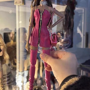 Könnte beinhalten: Eine Modepuppe mit langen braunen Haaren, die ein leuchtend rosa Kleid mit goldenem Reißverschluss, passende Oberschenkelstiefel und eine kleine Handtasche trägt. Die Puppe hat auch rosa Creolen und eine silberne Halskette.