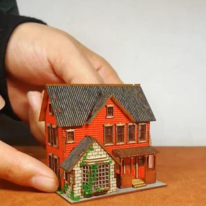 May include: A miniature wooden house model with a red exterior and a gray shingled roof. The model features a small porch, multiple windows, and a bay window with greenery. The house is held by a person.
