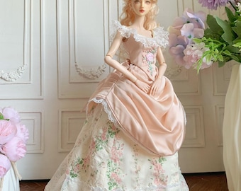 May include: A porcelain doll in a vintage-style gown. The dress is a soft pink with a floral patterned skirt and lace trim. The doll has blonde hair and is posed in front of a white wall with floral accents.