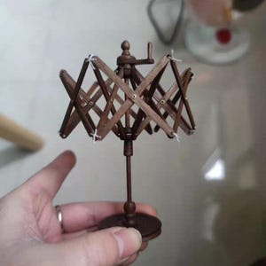 May include: A small, wooden yarn swift, featuring a collapsible design. The swift is made of dark brown wood and has a circular base. White thread secures the wooden arms. A hand crank is visible on the top.