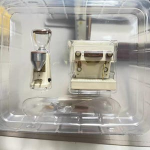 Puede incluir: Juego de cafetera y molinillo en miniatura en un recipiente de plástico transparente. La cafetera es de color blanco roto con detalles marrones, y el molinillo tiene una tolva transparente. El conjunto está diseñado para parecerse a una configuración de café real.