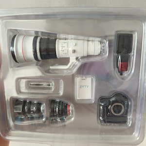 Pode incluir: Um kit de câmera em miniatura em uma bandeja de plástico transparente contendo uma teleobjetiva branca com um anel vermelho, um corpo de câmera preto, um flash preto, um pequeno cartão branco retangular com o texto ZYTV e duas lentes pretas.