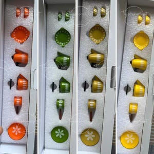 Könnte beinhalten: Ein Set bunter Vintage-Küchenspielzeug-Accessoires, darunter Teller, Knöpfe und Utensilien, angeordnet in einer Box. Die Teile sind in Orange, Grün und Gelb gehalten, mit weißen Akzenten. Das Set scheint ein Retro-Spielzeug zu sein.