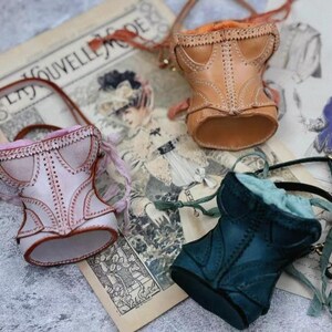 May include: Three miniature leather corset bags in pink, tan, and teal, each with a handle and decorative stitching. The bags are arranged on a vintage paper background with illustrations and text.