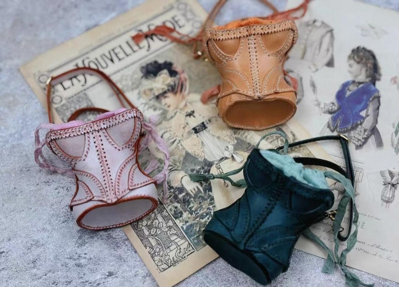May include: Three miniature leather corset bags in pink, tan, and teal, each with a handle and decorative stitching. The bags are arranged on a vintage paper background with illustrations and text.