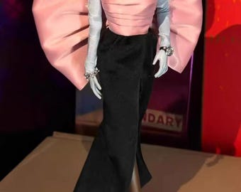 May include: A fashion doll in a pink strapless top with large bow details and a black skirt. The doll is accessorised with silver jewellery and long white gloves. The doll is wearing pink high heels.
