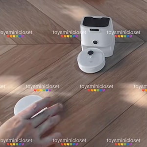 May include: A white, compact robotic vacuum cleaner with a square top and a circular base. The device is shown on a wooden floor, with a hand reaching towards the base. The product is designed for home cleaning.