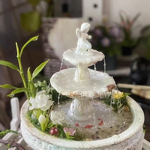 May include: A miniature white ceramic fountain with a tiered design, featuring a small angel figurine at the top. The fountain has water cascading down, surrounded by miniature flowers, greenery, and small red fish. The fountain is in a decorative, light-colored basin.