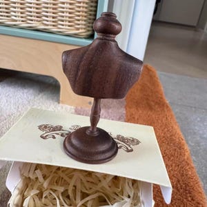 以下が含まれることがあります： 小さな濃い茶色の木製バストが台座にあり、おそらくジュエリーや小さなアクセサリーの展示用です。 バストは、紙の詰め物が入った装飾的な箱に表示されます。 箱には装飾的なデザインのクリーム色の蓋が付いています。