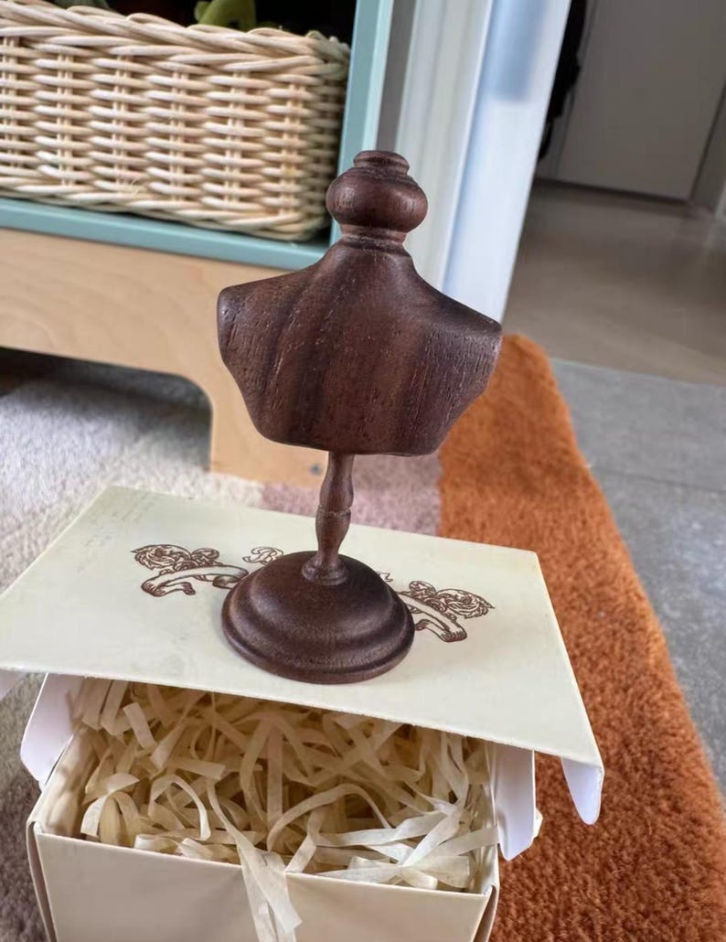 以下が含まれることがあります： 小さな濃い茶色の木製バストが台座にあり、おそらくジュエリーや小さなアクセサリーの展示用です。 バストは、紙の詰め物が入った装飾的な箱に表示されます。 箱には装飾的なデザインのクリーム色の蓋が付いています。