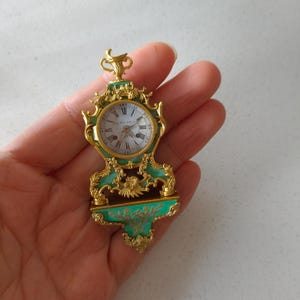 Könnte beinhalten: Eine Miniatur-Antiquitätenuhr mit goldfarbenem Rahmen und türkisfarbenem Sockel. Das Zifferblatt hat römische Ziffern und die Worte "VALERIA BALLE" sind auf das Zifferblatt gedruckt. Die Uhr hat eine verzierte Detaillierung und eine dekorative Oberseite.
