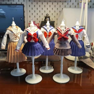 Puede incluir: Cinco uniformes escolares en miniatura sobre maniquíes blancos. Los uniformes presentan cuellos de marinero, faldas y lazos en varios colores, incluyendo azul, rojo y marrón. Los conjuntos se exhiben sobre una superficie de madera oscura.