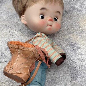 May include: A handcrafted doll with a large head, rosy cheeks, and brown hair. The doll wears a striped shirt, blue jeans, and brown leather boots. A brown leather bag with a strap is slung over its shoulder.
