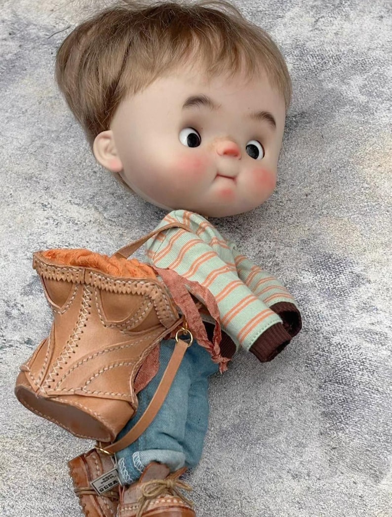 May include: A handcrafted doll with a large head, rosy cheeks, and brown hair. The doll wears a striped shirt, blue jeans, and brown leather boots. A brown leather bag with a strap is slung over its shoulder.
