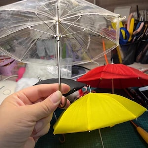 Puede incluir: Una colección de paraguas en miniatura de varios colores, incluyendo transparente, rojo, amarillo y negro. El paraguas transparente se sostiene en una mano, mostrando su cubierta transparente y su armazón plateado. Los otros paraguas están sobre una esterilla de corte verde.