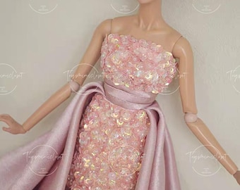 May include: A fashion doll wearing a strapless, short pink dress embellished with sequins. The dress is paired with a matching long, flowing overskirt. The doll has dark hair styled in an updo and is wearing pearl earrings.