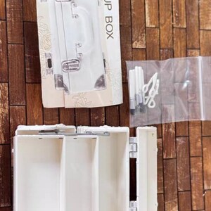 May include: A white makeup box with the words "MAKE UP BOX" printed on the packaging. The box is open, revealing several compartments. A clear plastic bag contains makeup tools. The background is a wooden surface.