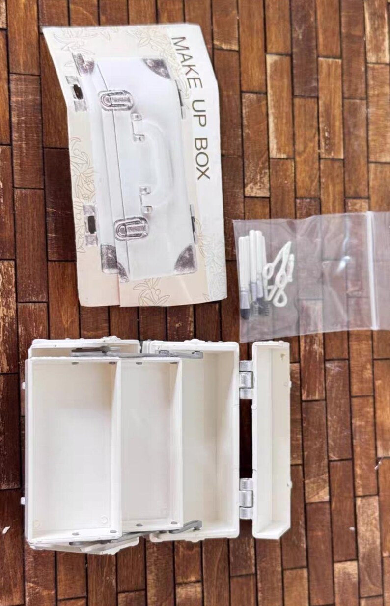 May include: A white makeup box with the words "MAKE UP BOX" printed on the packaging. The box is open, revealing several compartments. A clear plastic bag contains makeup tools. The background is a wooden surface.