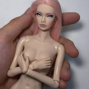 May include: A nude-colored fashion doll with articulated joints and long pink hair. The doll has detailed facial features, including blue eyes, long eyelashes, and pink lips. The doll is held by a hand.