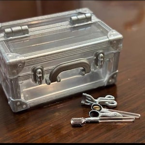 May include: A miniature silver-colored suitcase with a clear top, handle, and corner protectors. Beside it are miniature silver scissors and a comb, likely for a doll or craft project. The items are on a dark brown surface.