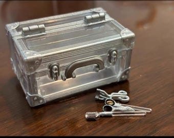 Könnte beinhalten: Ein kleiner silberfarbener Koffer mit transparentem Deckel, Griff und Eckschützern. Daneben befinden sich Miniatur-Silberscheren und ein Kamm, wahrscheinlich für eine Puppe oder ein Bastelprojekt. Auf einer dunkelbraunen Oberfläche.