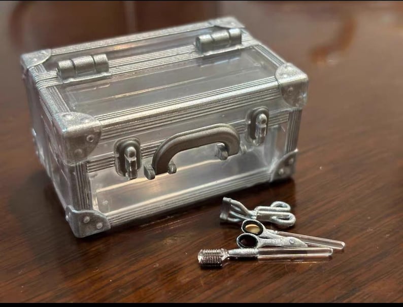 May include: A miniature silver-colored suitcase with a clear top, handle, and corner protectors. Beside it are miniature silver scissors and a comb, likely for a doll or craft project. The items are on a dark brown surface.