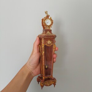 May include: A miniature, ornate clock with a brown and gold finish. The clock has a small, round face and is held in a hand. The clock is approximately 6 inches tall.