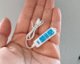 May include: A miniature white power strip with three blue outlets and a white cord. The power strip is held in a hand, showcasing its small size and detailed design. The outlets have a blue border.