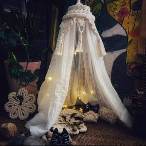 May include: A cream-coloured macrame canopy with a layered design, featuring tassels and fairy lights. The canopy drapes over a small area, creating a cosy atmosphere. Decorative macrame coasters and a black cat-shaped item are in the foreground.