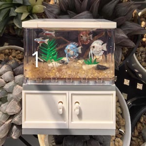 Könnte beinhalten: Ein Miniatur-Aquarium mit bunten Spielzeugfischen, grünen Pflanzen und dekorativem Kies. Das transparente Becken steht auf einem weißen Schrank mit zwei Türen. Die Zahl "1" ist auf der Vorderseite des Beckens zu sehen.