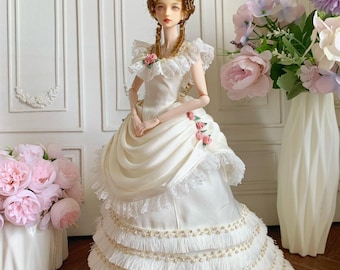 May include: A porcelain doll in a white gown with a high, powdered wig. The dress features lace trim, pink floral accents, and tiered skirt with fringe and gold beading. The doll is posed in front of a white wall with decorative panels.