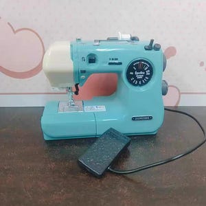 Può includere: Una macchina da cucire Janome turchese con la parte superiore color crema e un pedale nero. La macchina ha un quadrante circolare con opzioni di selezione dei punti e il testo "Épolku". La macchina è su una superficie marrone.