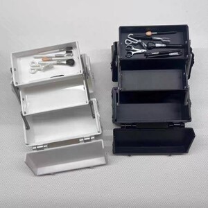 May include: Two miniature makeup cases, one white and one black, are open to reveal their contents. The white case holds brushes and scissors, while the black case contains brushes, scissors, and other tools. The cases are on a light gray surface.