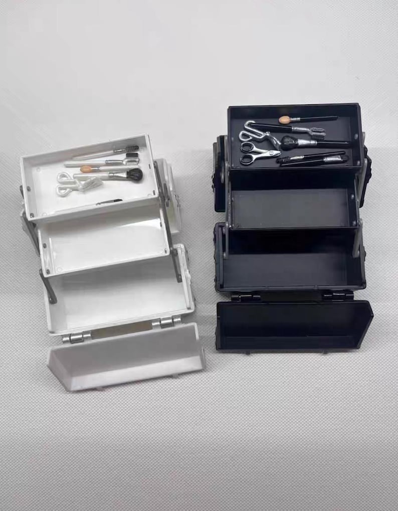 May include: Two miniature makeup cases, one white and one black, are open to reveal their contents. The white case holds brushes and scissors, while the black case contains brushes, scissors, and other tools. The cases are on a light gray surface.