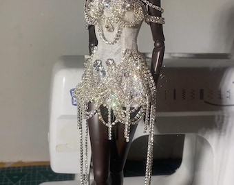 May include: A dark-skinned doll wearing a white, embellished dress. The dress is adorned with sparkling rhinestones and dangling chains. The doll is posed in front of a sewing machine, suggesting a fashion or craft theme.
