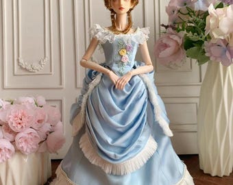 May include: A doll in a light blue satin gown with white fringe and gold trim. The doll has a brown wig and is wearing a necklace. The dress has a ruffled skirt and is decorated with embroidered flowers.