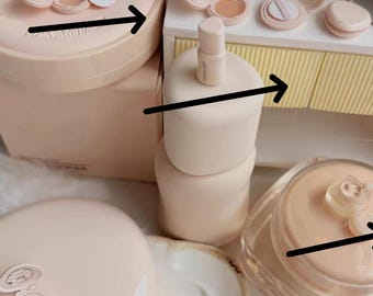 Könnte beinhalten: Eine Sammlung hellrosa Kosmetikprodukte, darunter Kompakte, Flaschen und Tiegel. Die Artikel sind auf einer weißen Oberfläche angeordnet, mit einem kleinen dekorativen Blumenarrangement im Hintergrund. Die Gesamtästhetik ist weich und feminin.
