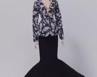 May include: A doll in a black and white butterfly-print jacket with a low neckline and a long black skirt. The skirt has a flared, fishtail design. The doll has dark hair and red lipstick.