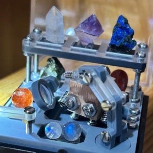 Puede incluir: Una exhibición con varios cristales y piedras preciosas. La disposición incluye una punta de cuarzo transparente, un cristal de fluorita púrpura y un espécimen de azurita azul. También hay piedras adicionales y una lupa.